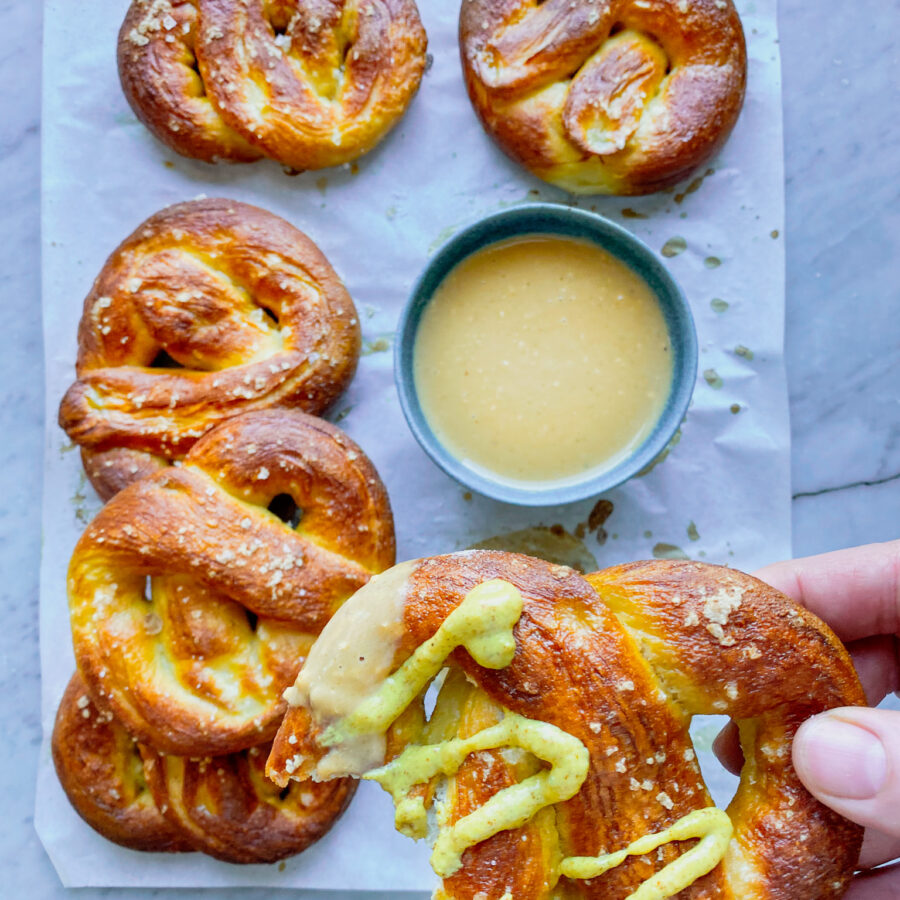 5 soft pretzels and cheese sauce and one pretzel with mustard and a bite taken out of it being held by a hand