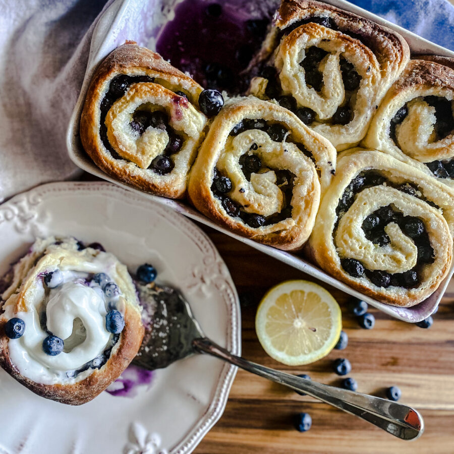 blueberry sweet rolls with one served on a plate with frosting and lemons and blueberries
