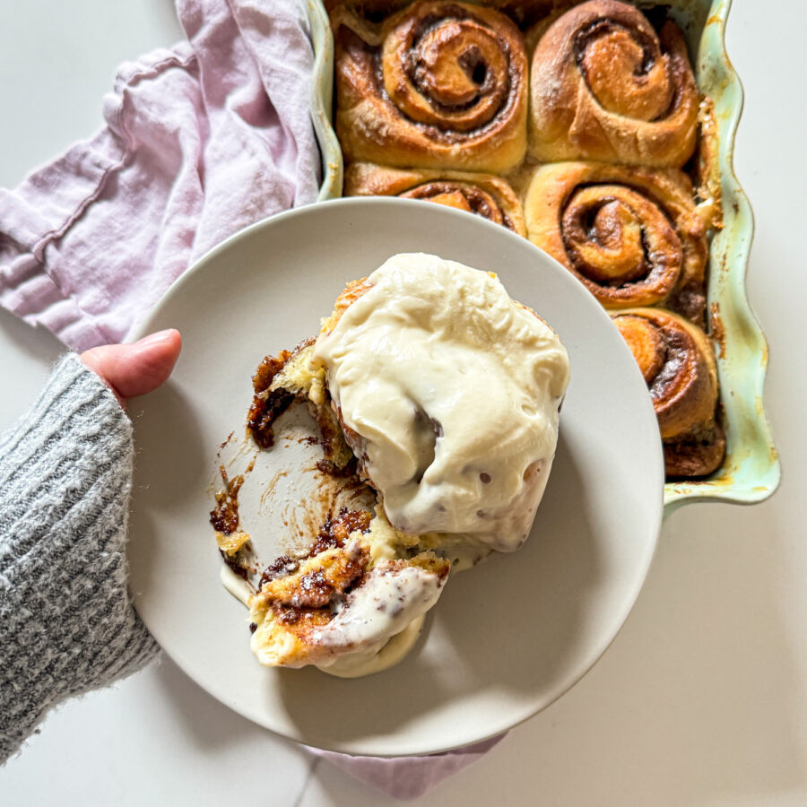 Hand with a sweather holding a plate with a frosted cinnamon roll and a pan of cinnamon rolls