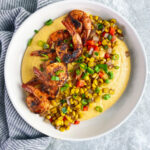 white bowl with yellow cheese grits, blackened shrimp, and sautéd onions, peppers and corn.