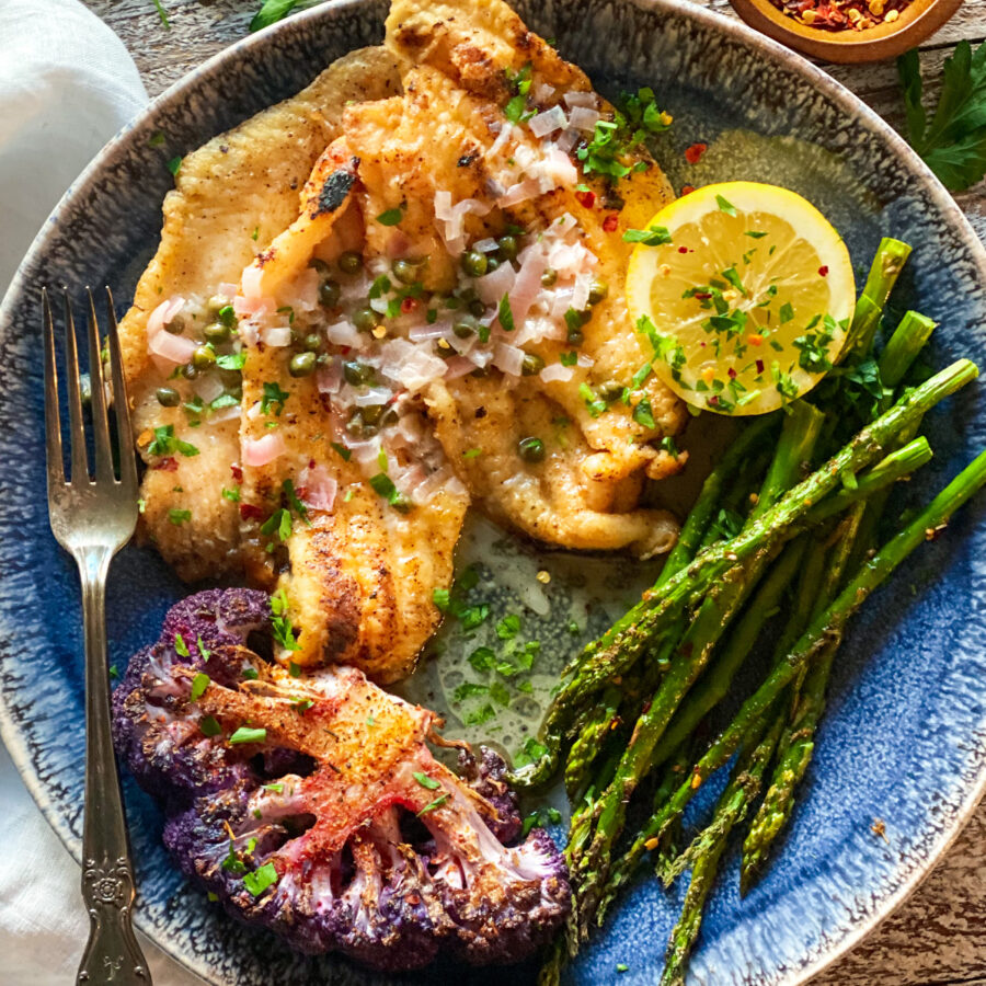 Dover Sole with white wine sauce on a blue plate with purple cauliflower and asparagus and a lemon