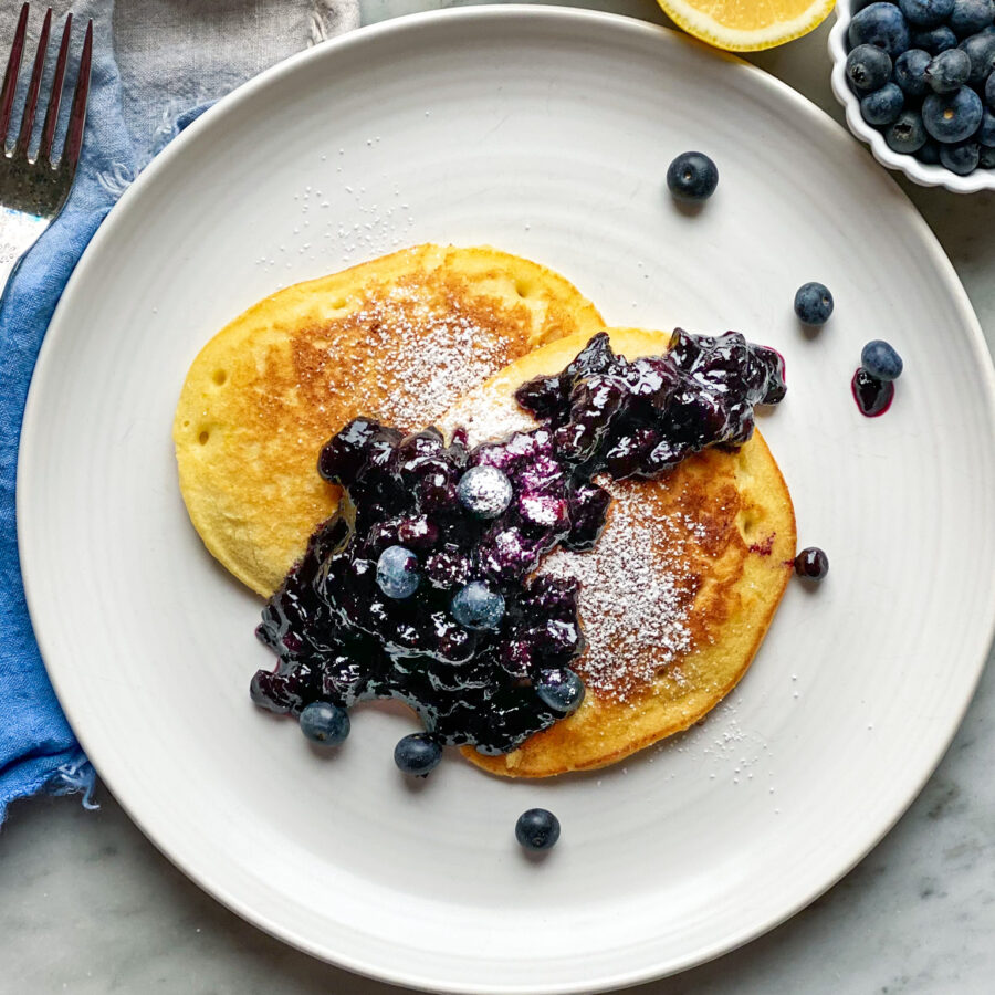 2 lemon pancakes on a plate with blueberry compote and a sliced lemon and blueberries