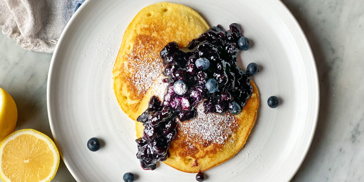 2 lemon pancakes on a plate with blueberry compote and a sliced lemon and blueberries