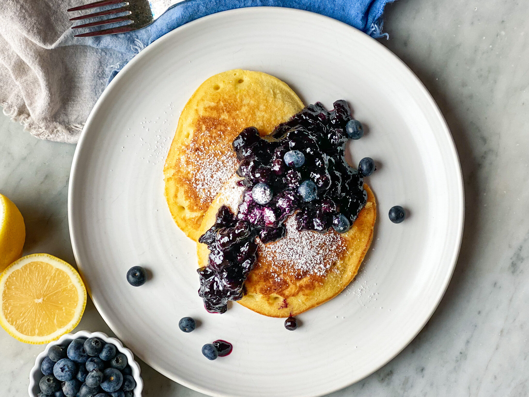 2 lemon pancakes on a plate with blueberry compote and a sliced lemon and blueberries