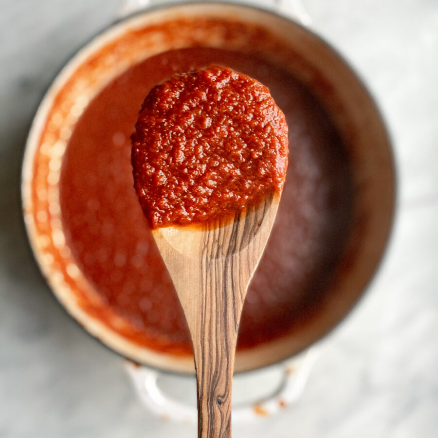 A wooden spoon with tomato sauce and a pot of tomato sauce