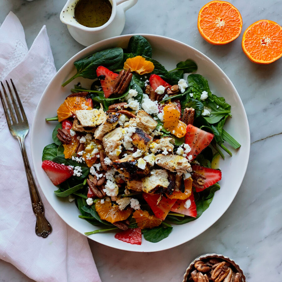 Spinach salad with oranges, strawberries, chicken, pecans, and feta, with a bowl of strawberries and pecans and an orange and a pitcher of poppyseed dressing