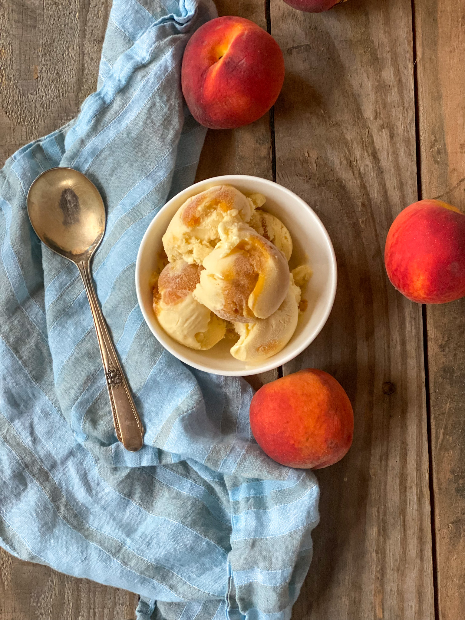 Peach ice cream in a bowl with peaches and a blue napkin and a spoon