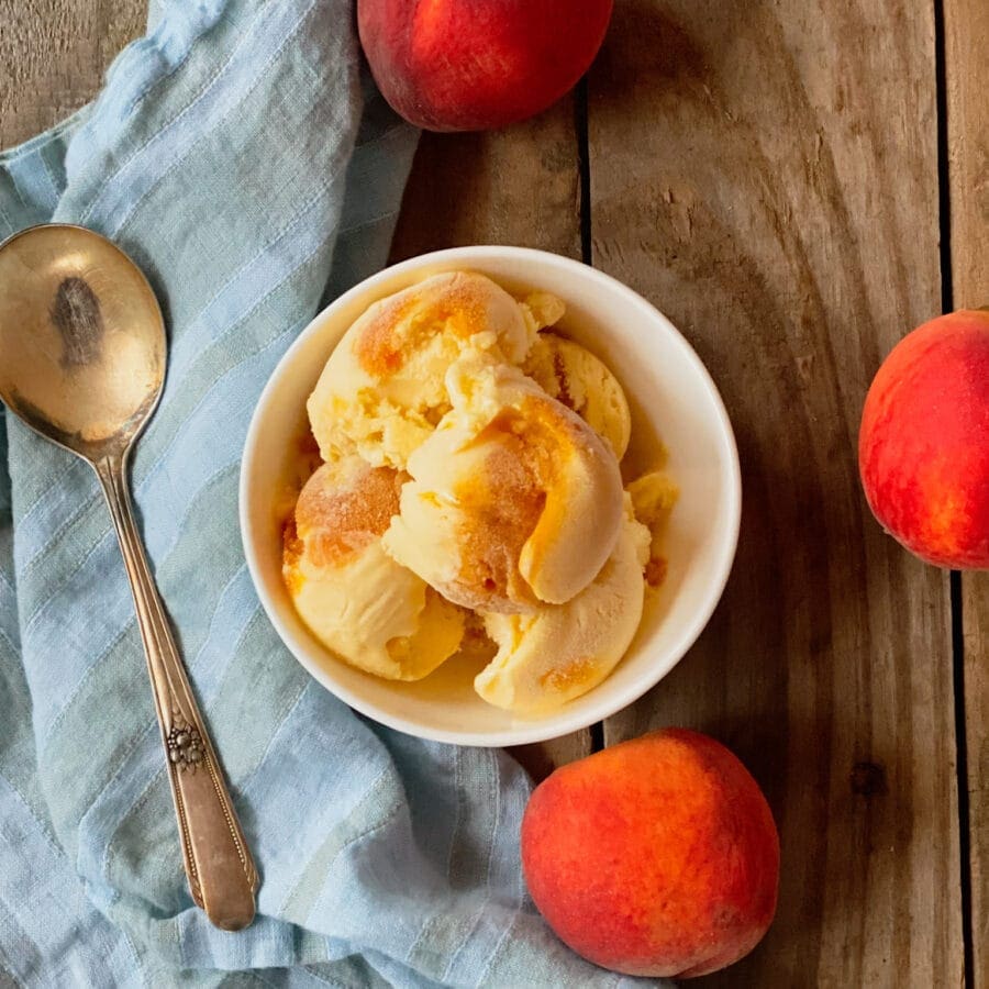 peach icecream in a bowl with a silver spoon on a bue stripe napkin with 3 peaches