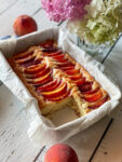 Peach topped cake with a slice out and peaches and flowers nearby