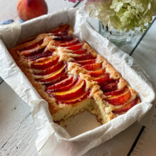 Peach topped cake with a slice out and peaches and flowers nearby