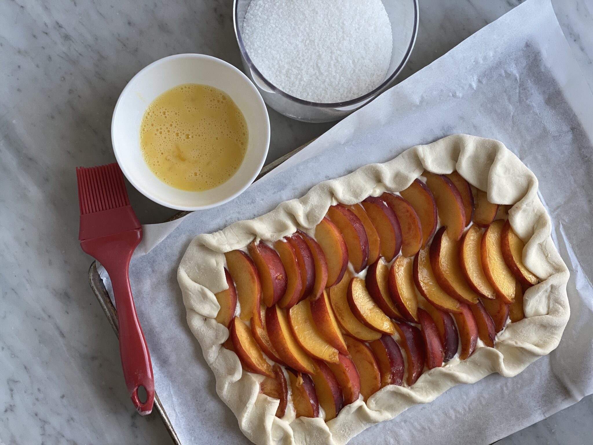 Whisked egg in a bowl, course sugar in a bowl, and peach galette, unbaked