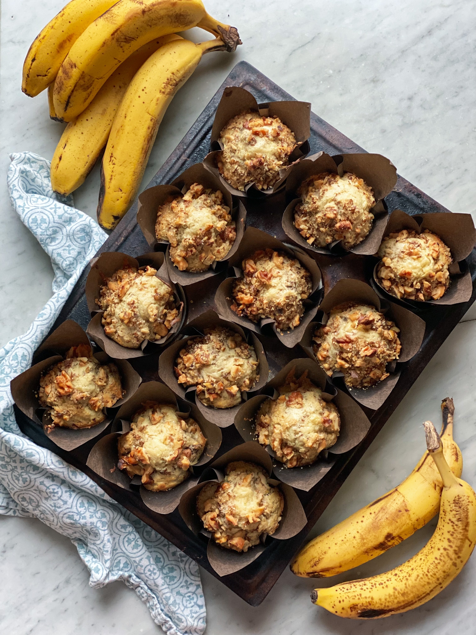 a dozen banana nut muffins in a muffin pan with a napkin and bananas laying next to them