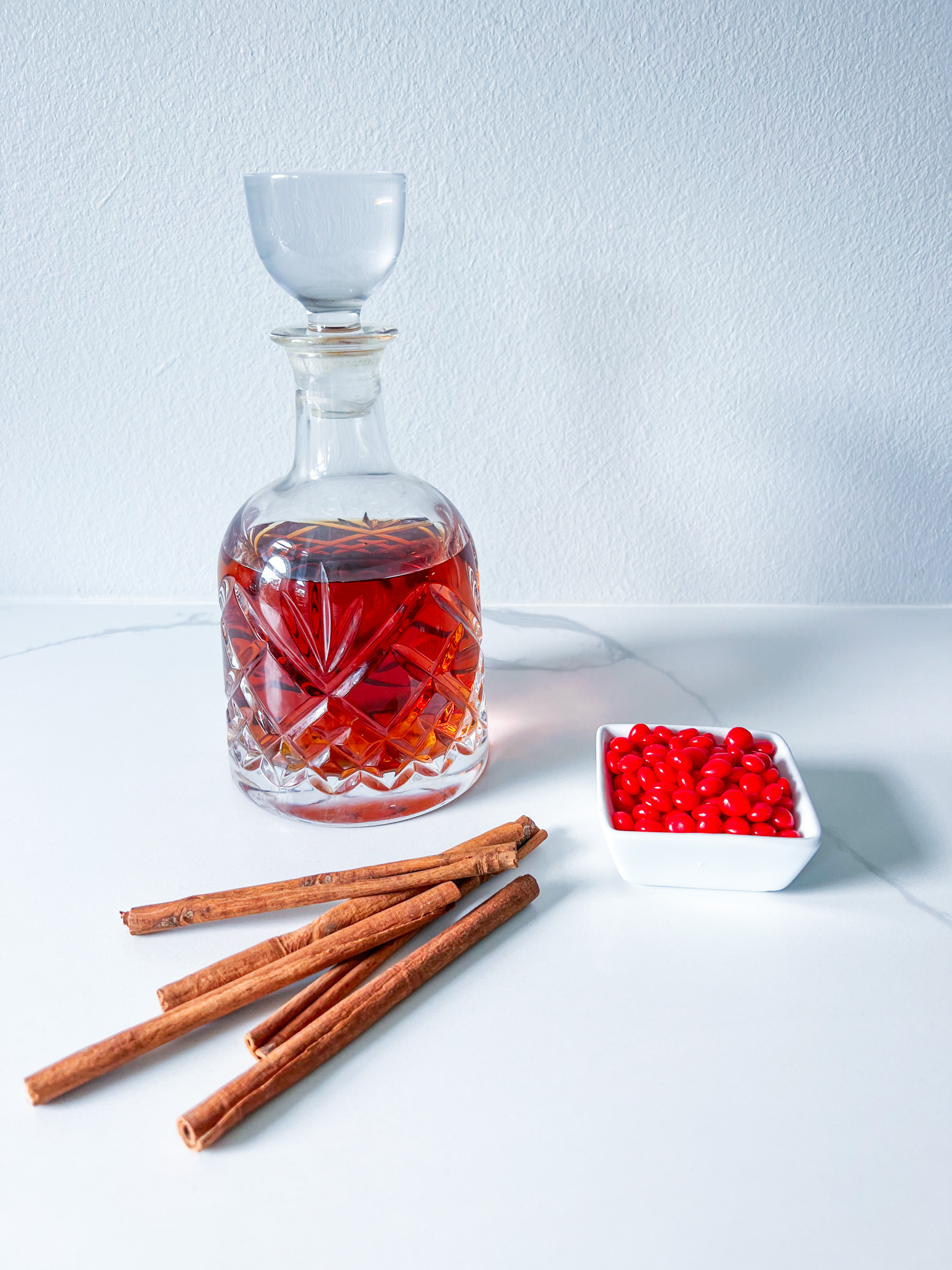 a bottle of bourbon in a decanter with a lid, long cinnamon sticks and a square bowl of red hot candies