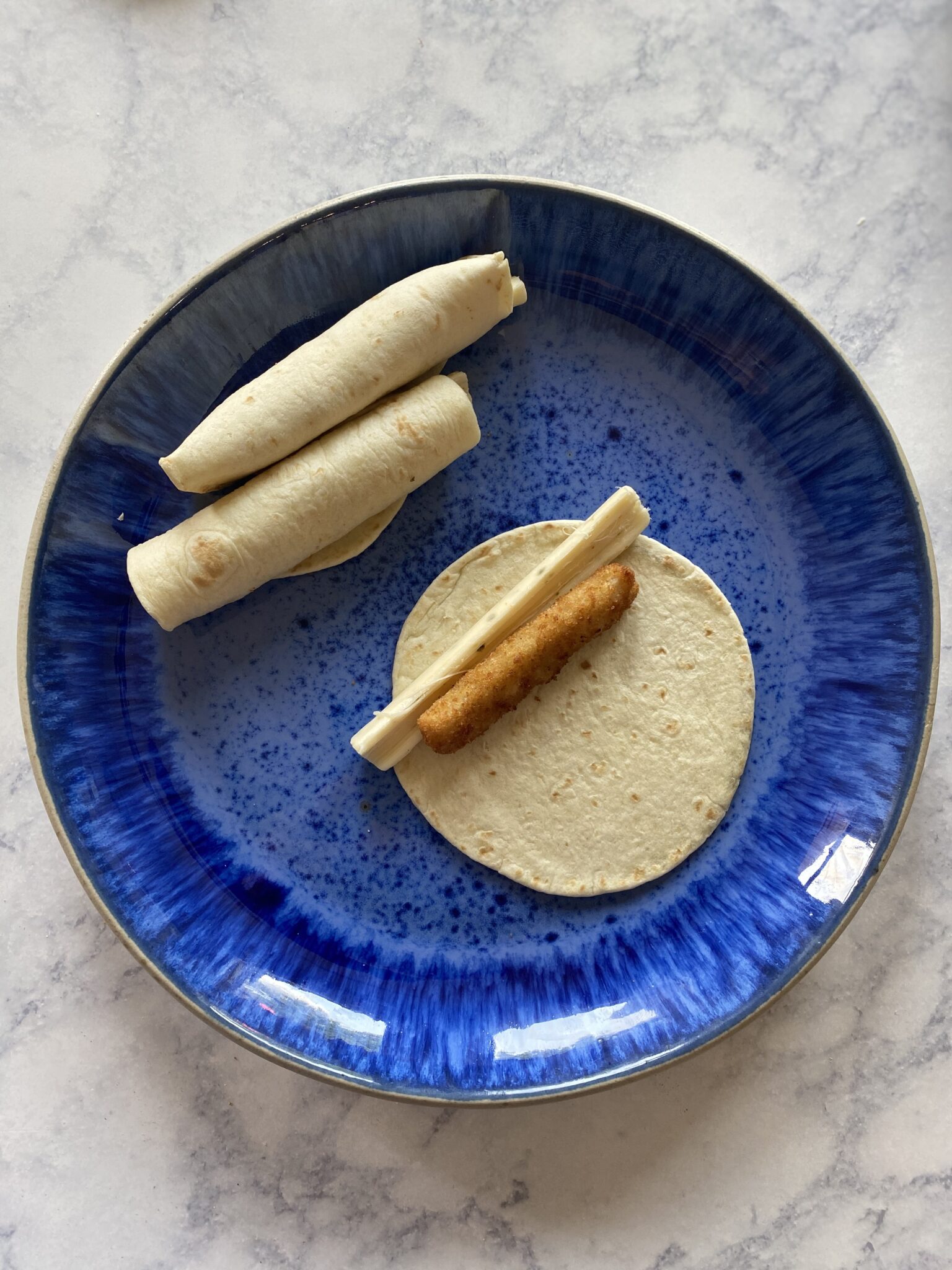 Fish stick and cheese stick in a tortilla with 2 rolled up, on a blue plate