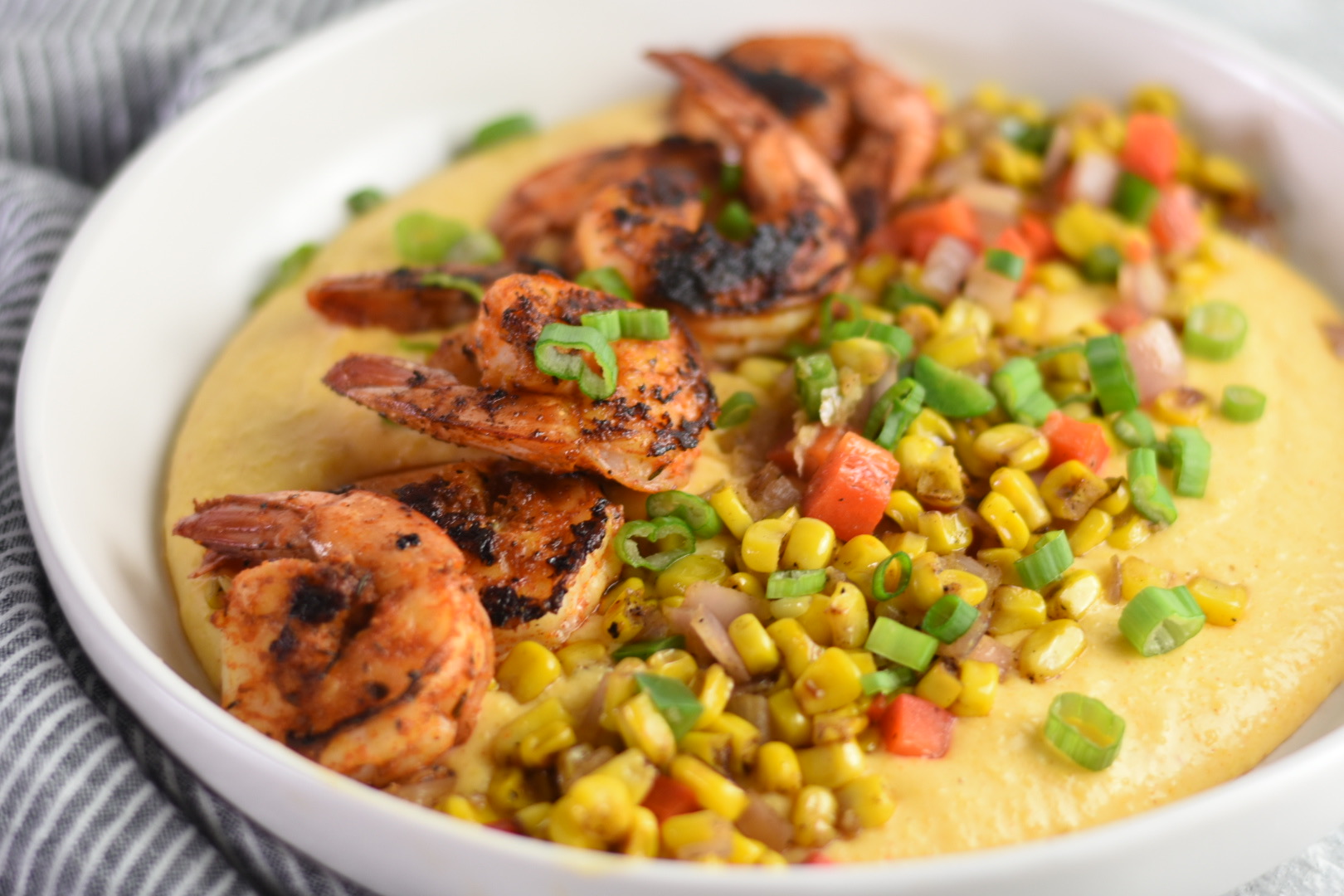 A white bowl filled with creamy yellow grits, topped with grilled shrimp, vibrant corn, chopped bell peppers, diced onions, and garnished with chopped green onions. The dish is placed on a grey and white striped cloth.