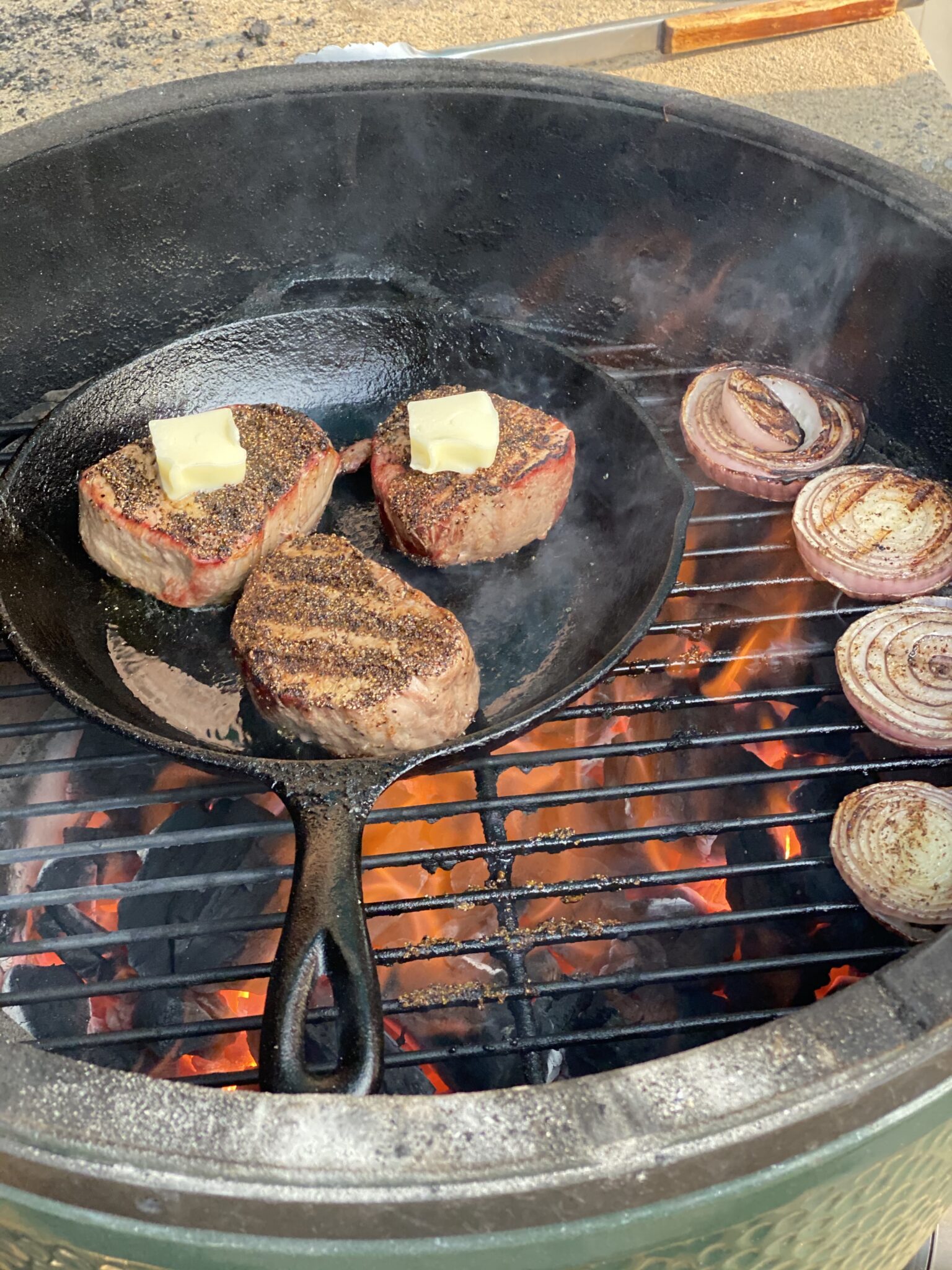 steaks in a cast iron skillet with butter on them on a grill with fire and onions
