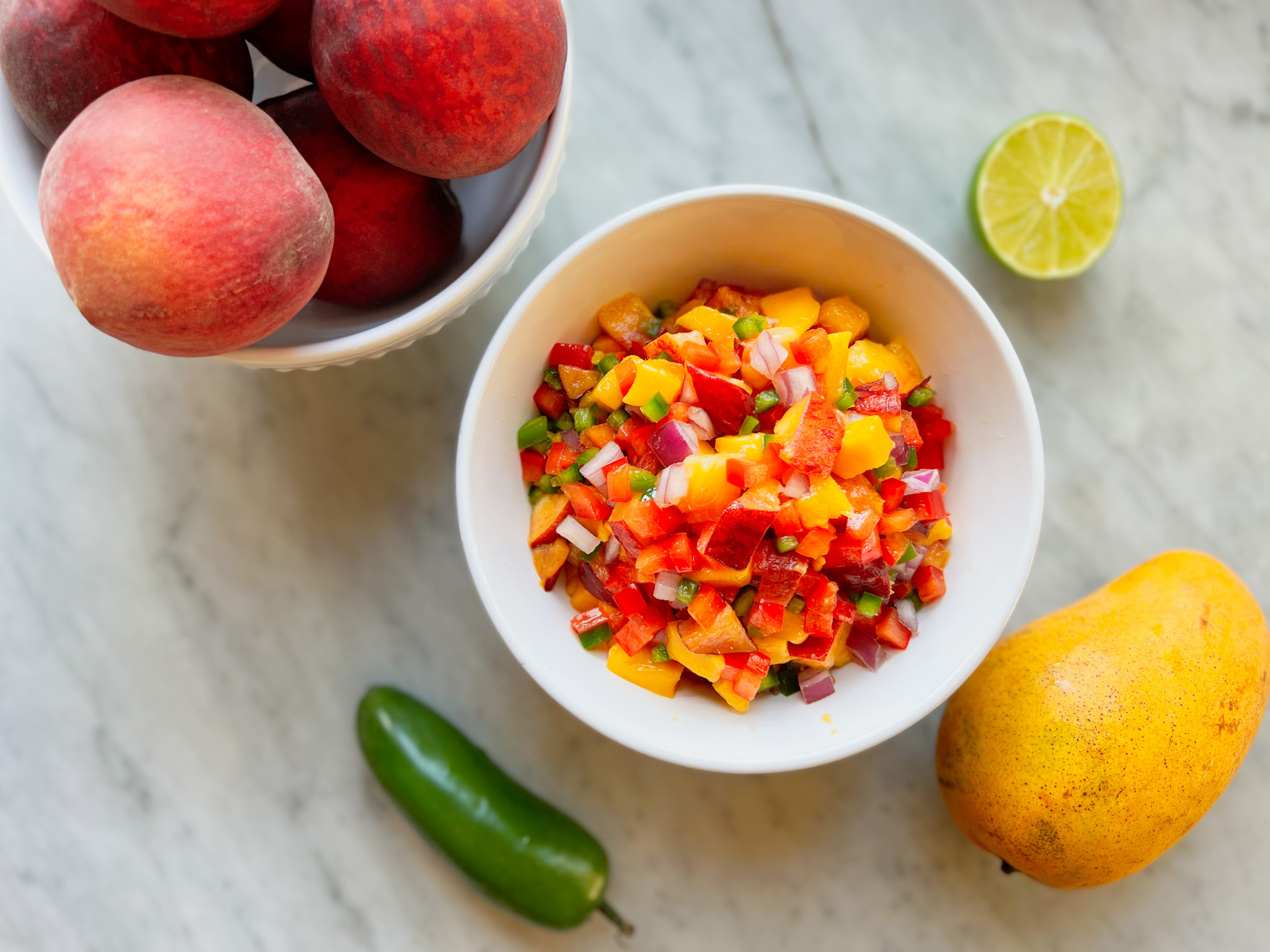 Mango peach salsa in a bowl with a bowl of peaches, jalapeño, mango and lime