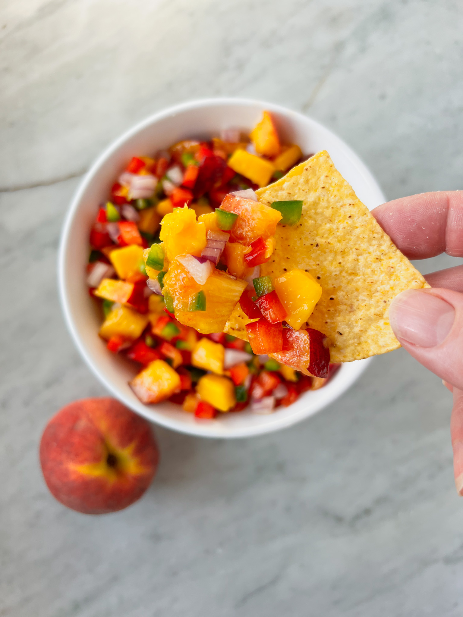 a peach, a white bowl with mango-peach salsa and a hand with a chip and salsa