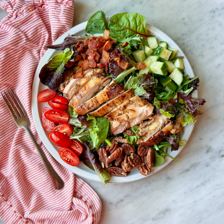 sliced smoked grilled chicken thighs on a salad with tomatoes, pecans, candied bacon, and cucumbers