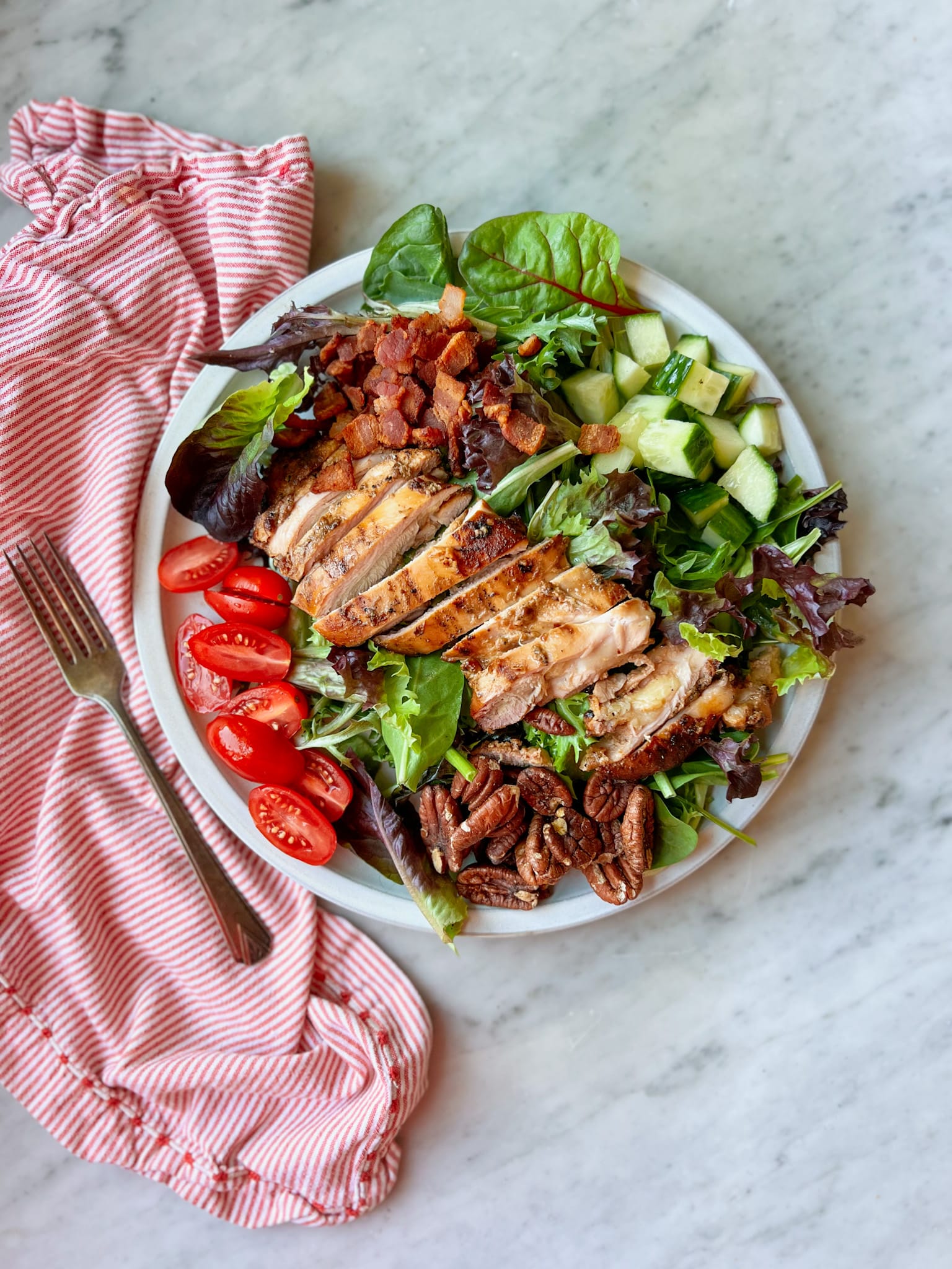 Grilled chicken thighs on a salad with tomatoes, cucumber, bacon, pecans and lettuce.  