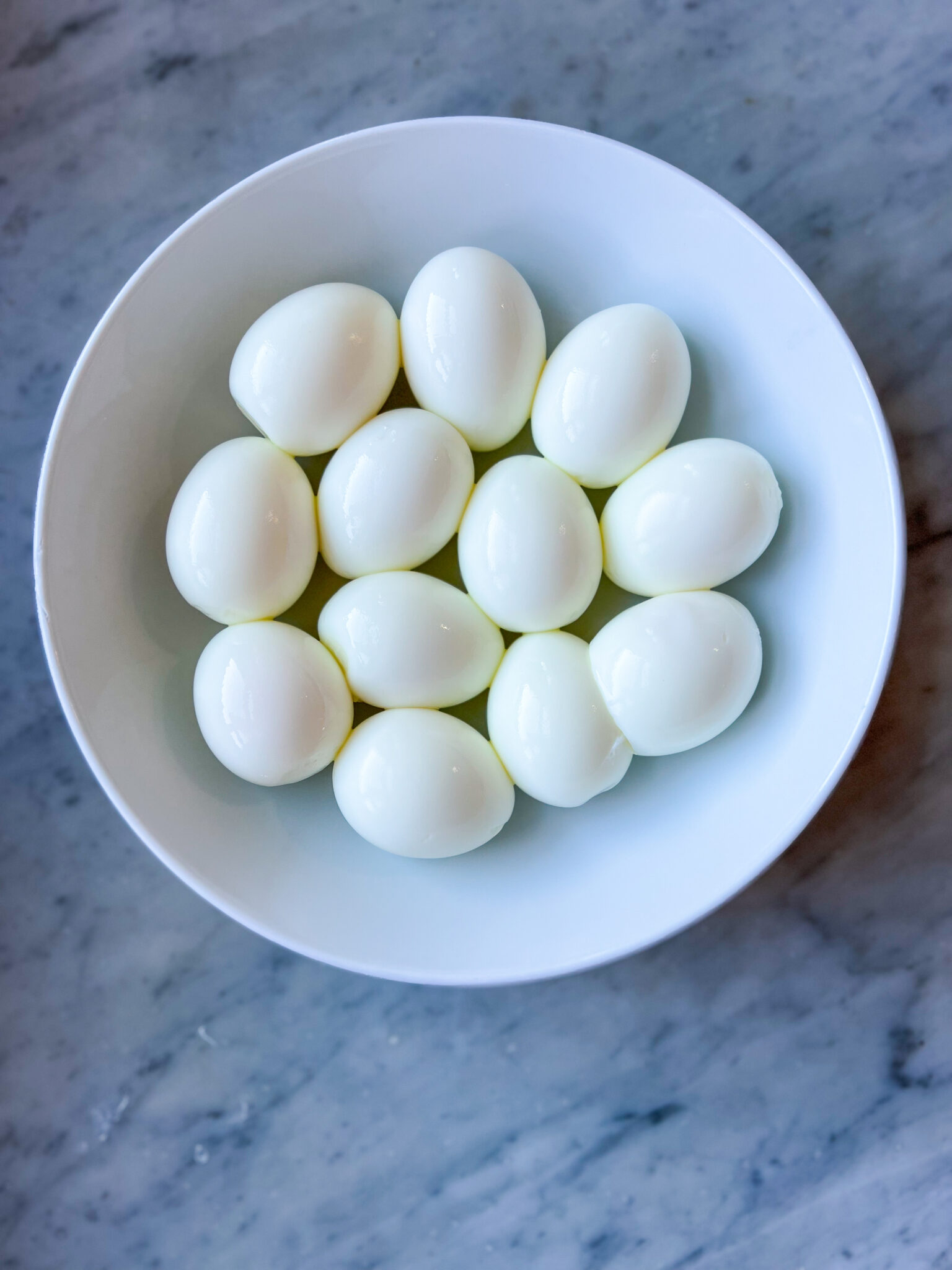 a dozen hard boiled eggs in a white bowl on a marble table