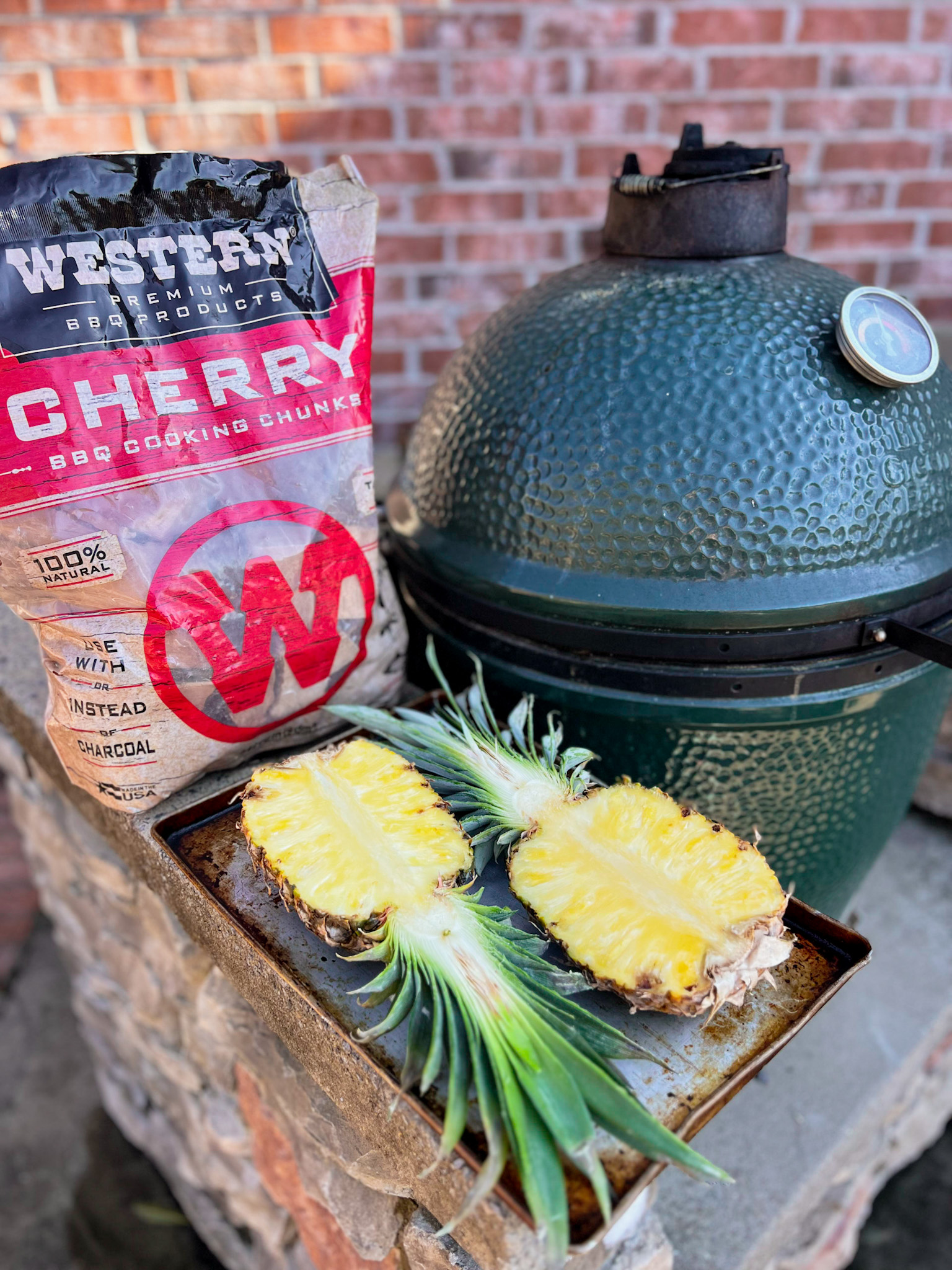 wetern wood bbq cherry wood chunks in a bag and a whole pineapple cut in half sitting next to a big green egg smoker