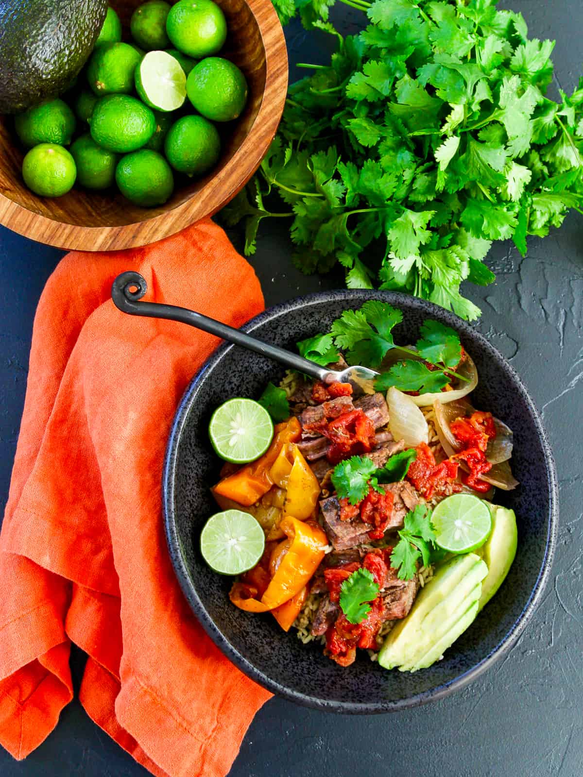 Slow cooker shredded beef in a bowl with limes and cilantro and an orange napkin
