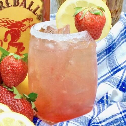 Strawberry Spiked Lemonade with Fireball Whisky garnished with a lemon and a strawbery with a blue and white napkin and a bottle of fireball