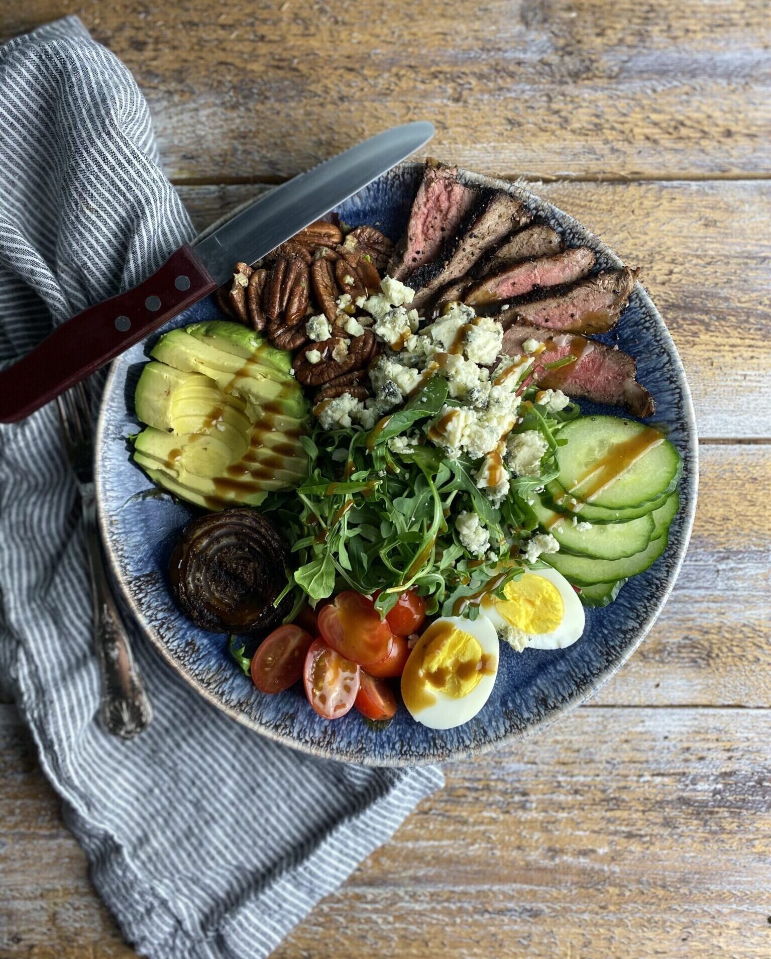 steak salad with bleu cheese, roasted pecans, cucumber, eggs, avocado in a bowl with a knife