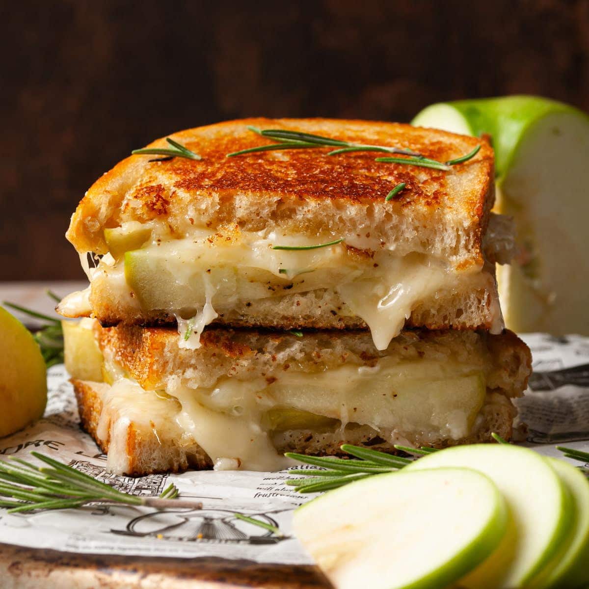 a stuffed brie and apple grilled cheese sliced in half and stacked, with green apple slices on a plate with a sprinkle of rosemary