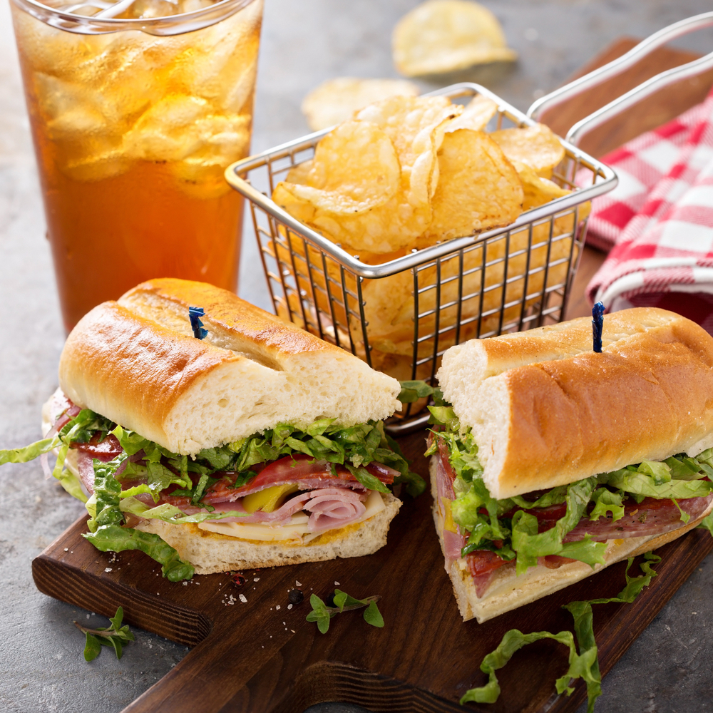 a grinder sandwich cut in half on a board with potato chips and iced tea