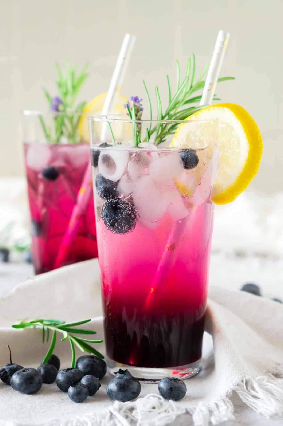 Two tall glasses of blueberry lemonade with rosemary and lemon garnish and straws
